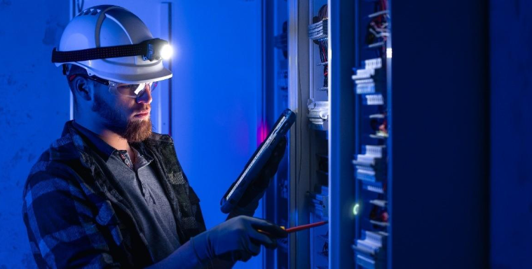 Technician in a hard hat using a tablet to inspect electrical equipment.